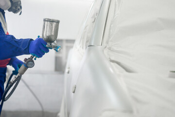 A mechanic is using a blue spray gun to paint a car. A car painter wearing blue uniform and gloves uses a spray gun in a car drying room.