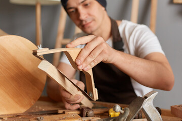 Closeup portrait of handsome young adult man carpenter wearing white T-shirt and brown apron sanding wooden wooden block, making wooden chair, likes his hobby in carpentry workshop.