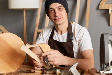 Portrait of smiling attractive young adult man carpenter dressed brown apron and black cap, sanding wooden wooden block in his workshop, looking at camera.