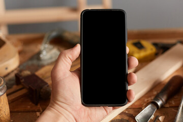 Closeup of mobile phone with blank black scree in man's hand on background of carpentry tools and...