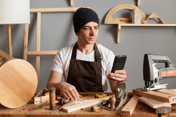 Young adult man carpenter wearing a black hat and brown apron, sitting at the table of his...