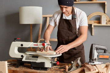Horizontal shot of concentrated man carpenter wearing brown apron and black cap working in his joinery, works with an electric jigsaw, making wood products.