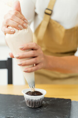 Decorate the cake with white cream. Using a pastry bag, the pastry chef makes cupcakes. Female hands applying butter cream on delicious chocolate cupcakes, homemade baking