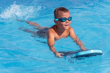 Happy boy child in swimming goggles with float foam board tool swim in pool in sunny day. Safe pool...