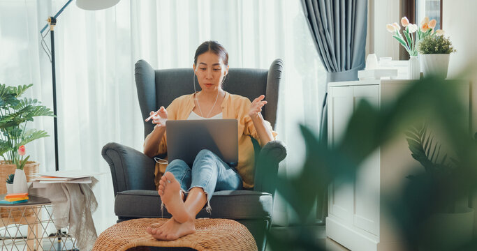 Young asia female girl or university student sit on sofa chair with computer laptop wear earphone look at screen enjoy online meeting presenting work in living room at house. Home lifestyle concept.