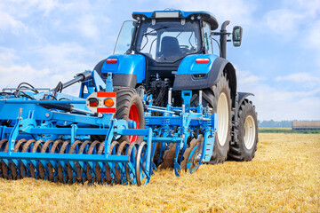 Fototapeta premium A blue agricultural tractor pulls a harrow across a mowed wheat field on a summer day.