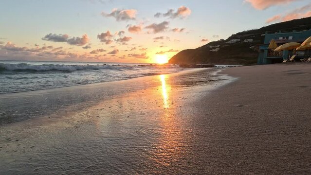 Golden setting sun off the point of land lights clouds and reflects off wet sandy beach as wave retreats back to the sea with reflection following.