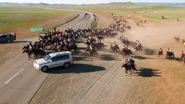 Close drone shot of a group of Mongolian jockeys training for a traditional Naadaam. Horses running in a fast canter crossing a highway in the desolate steppe landscape.
