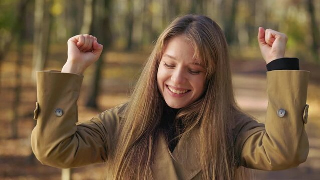 Front view of pretty lady standing in park, showing hurray, dancing. Beautiful woman wearing fashionable clothes, looking at camera, smiling. Concept of happiness and enjoyment.
