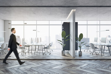 Side view of young businessman walking in modern glass office interior with wooden flooring, furniture, window with city view and other objects.