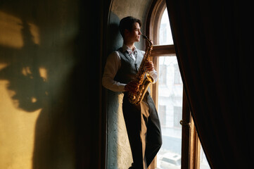Creative young man playing saxophone standing on windowsill