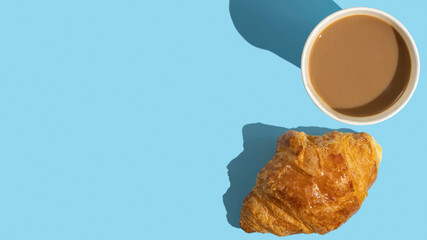 Coffee cup and fresh croissant for tasty breakfast on blue background with copy space.