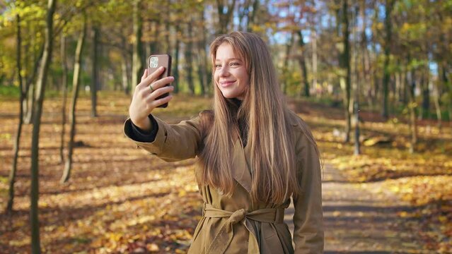 Side view of stylish lady wearing fashionable clothes, walking in park in autumn. Beautiful, young female taking selfie, photo, smiling. Concept of happiness and enjoyment.