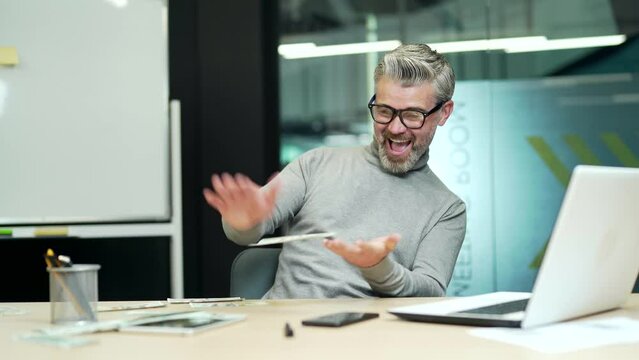 Cheerful mature gray haired bearded businessman happily scatters dollar bills at workplace in modern office. A happy entrepreneur rejoices at a good income and, laughing, frantically plays with money
