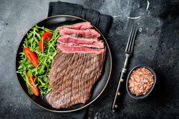 Grilled medium rare flank beef steak with salad in a plate. Black background. Top view
