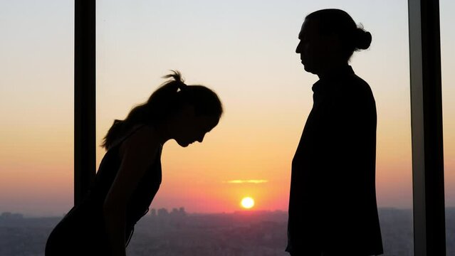 Woman and man say goodbye in Japanese style, making small bows. Silhouettes of two people standing in an office against window, sunset and dim cityscape seen on background