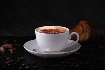 Italian breakfast on a black background. Cappuccino with croissant on a black background. White cup with cappuccino on a dark background, coffee beans on the table.