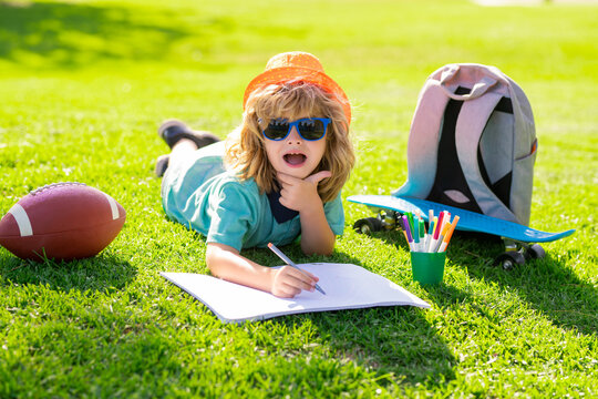 Little painter draw pictures outdoor. Clever school boy doing homework, writing on copy book in green grass of park. Back to school, knowledge, education, learning concepts.