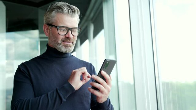 Mature gray haired bearded businessman in glasses uses smartphone while standing near window in modern office. Confident smiling entrepreneur checking messages on his mobile phone or chatting online