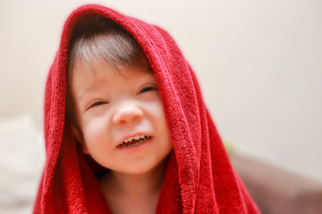 A laughing two-year-old boy with a broken tooth after a bath wrapped in a red towel at home