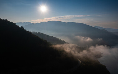 Aerial view of a foggy morning sunrise over a lake and a dam in the mountains next to Siriu Romania, with a cliff road winding
