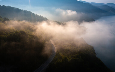 Aerial view with a road winding through a forest during a foggy morning sunrise and clouds of mist