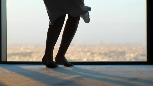 Emotional woman bouncing with joy, lower half shot. We can see her feet and the floor against the window. Blurred city panorama in the background.