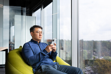 Serious thinking asian man reading news on phone, programmer using app on smartphone testing new program, businessman worker sitting on soft chair near window in modern office interior.