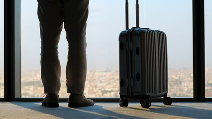 Tourist is nervous whether his flight has been cancelled. Man raise one leg and scratches his calf with foot. Packed suitcase stands next to him. Blurred cityscape seen in background, low half shot