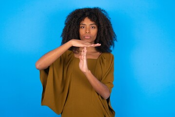 young woman with afro hairstyle wearing brown dress over blue wall being upset showing a timeout gesture, needs stop, asks time for rest after hard work,