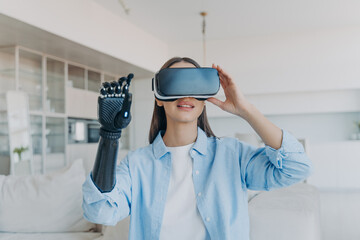 Disabled person gets rehabilitation. Handicapped girl with cyber arm in vr goggles at home.