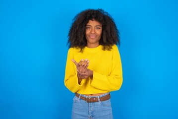 young woman with afro hairstyle wearing yellow sweater over blue wall Suffering pain on hands and fingers, arthritis inflammation