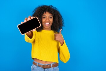 Portrait young woman with afro hairstyle wearing orange crop top over blue wall holding in hands cell showing giving black screen thumb up