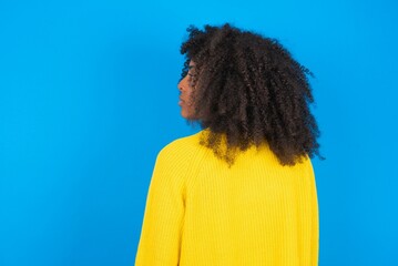 The back side view of a young woman with afro hairstyle wearing orange crop top over blue wall....