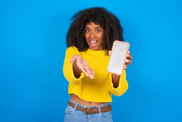 young woman with afro hairstyle wearing orange crop top over blue wall with a mobile. presenting smartphone. Advertisement concept.