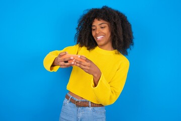 Portrait of an excited young woman with afro hairstyle wearing orange crop top over blue wall playing games on mobile phone.