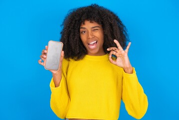 young woman with afro hairstyle wearing orange crop top over blue wall holding in hands cell...