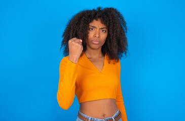 young woman with afro hairstyle wearing orange crop top over blue wall shows fist has annoyed face...
