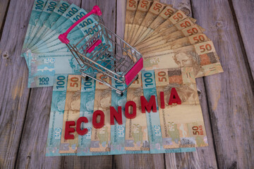 Brazilian banknotes. Banknotes of 50 and 100 reais in the background with the word "economia" in Portuguese (economy, in English), in red, with a mini supermarket cart. Selective focus.