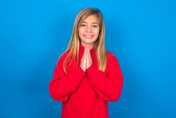 caucasian teen girl wearing red sweater over blue studio background keeps palms together, has pleased expression. Glad attractive male makes request, pleads for mercy. Hopeful young adult.