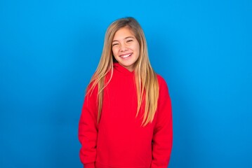 caucasian teen girl wearing red sweater over blue studio background with broad smile, shows white teeth, feeling confident rejoices having day off.