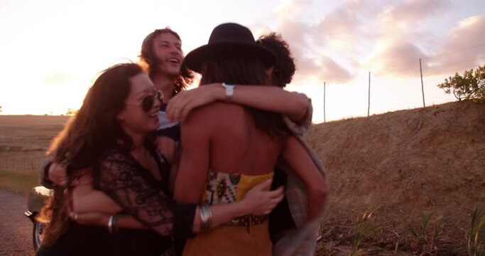 Group of teenager friends dressed in hipster style partying in a country road by cheering with raised arms and embracing with afternoon summer sun flare