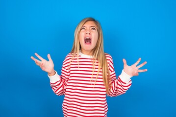 caucasian teen girl wearing striped shirt over blue studio background crying and screaming. Human...
