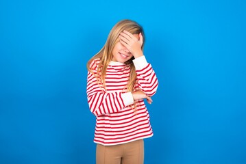 caucasian teen girl wearing striped shirt over blue studio background making facepalm gesture while smiling amazed with stupid situation.