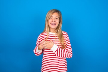 Honest caucasian teen girl wearing striped shirt over blue studio background keeps hands on chest, touched by compliment or makes promise, looks at camera with great pleasure.