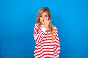 caucasian teen girl wearing striped shirt over blue studio background , holding his nose because of a bad smell.