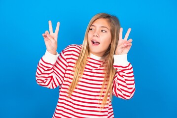 Isolated shot of cheerful caucasian teen girl wearing striped shirt over blue studio background makes peace or victory sign with both hands, feels cool.