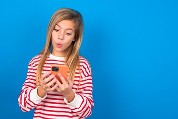 caucasian teen girl wearing striped shirt over blue studio background looks with bugged eyes, holds modern smart phone, receives unexpected message from friend, reads reminder.