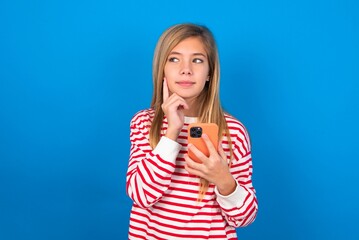 Thinking dreaming caucasian teen girl wearing striped shirt over blue studio background using mobile phone and holding hand on face. Taking decisions and social media concept.