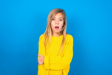 Shocked embarrassed caucasian teen girl wearing yellow sweater over blue studio background keeps...